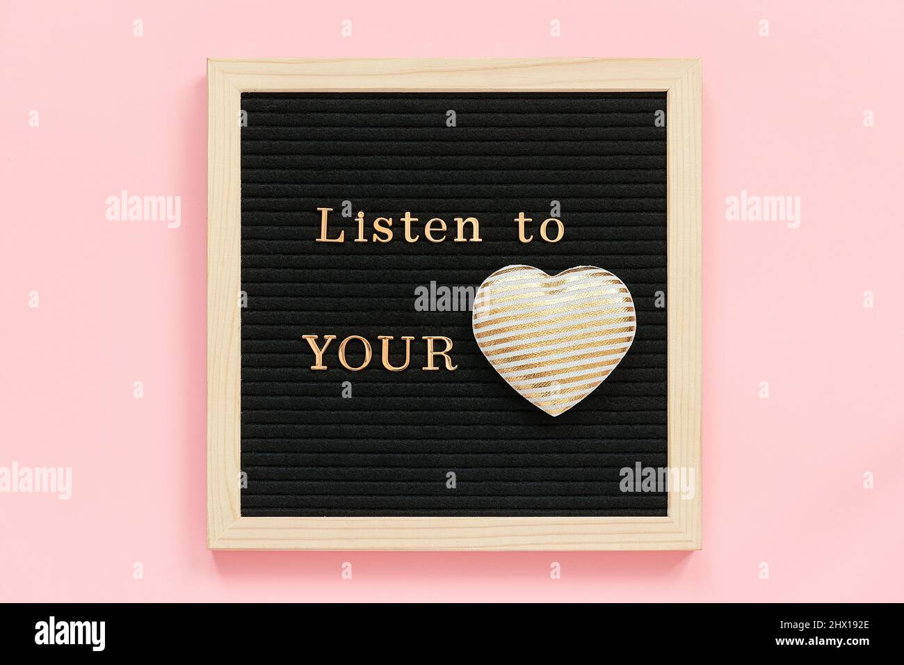 Ascoltate il vostro cuore. Citazione motivazionale in lettere d'oro e cuore tessile decorativo su bordo nero lettera su sfondo rosa. Vista dall'alto Conc. A disposizione piatta Foto Stock