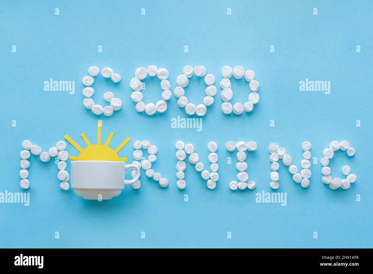 Testo "Buongiorno" da marshmallow e tazza di caffè con il sole giallo che sorge su sfondo blu. Concetto Foto Stock