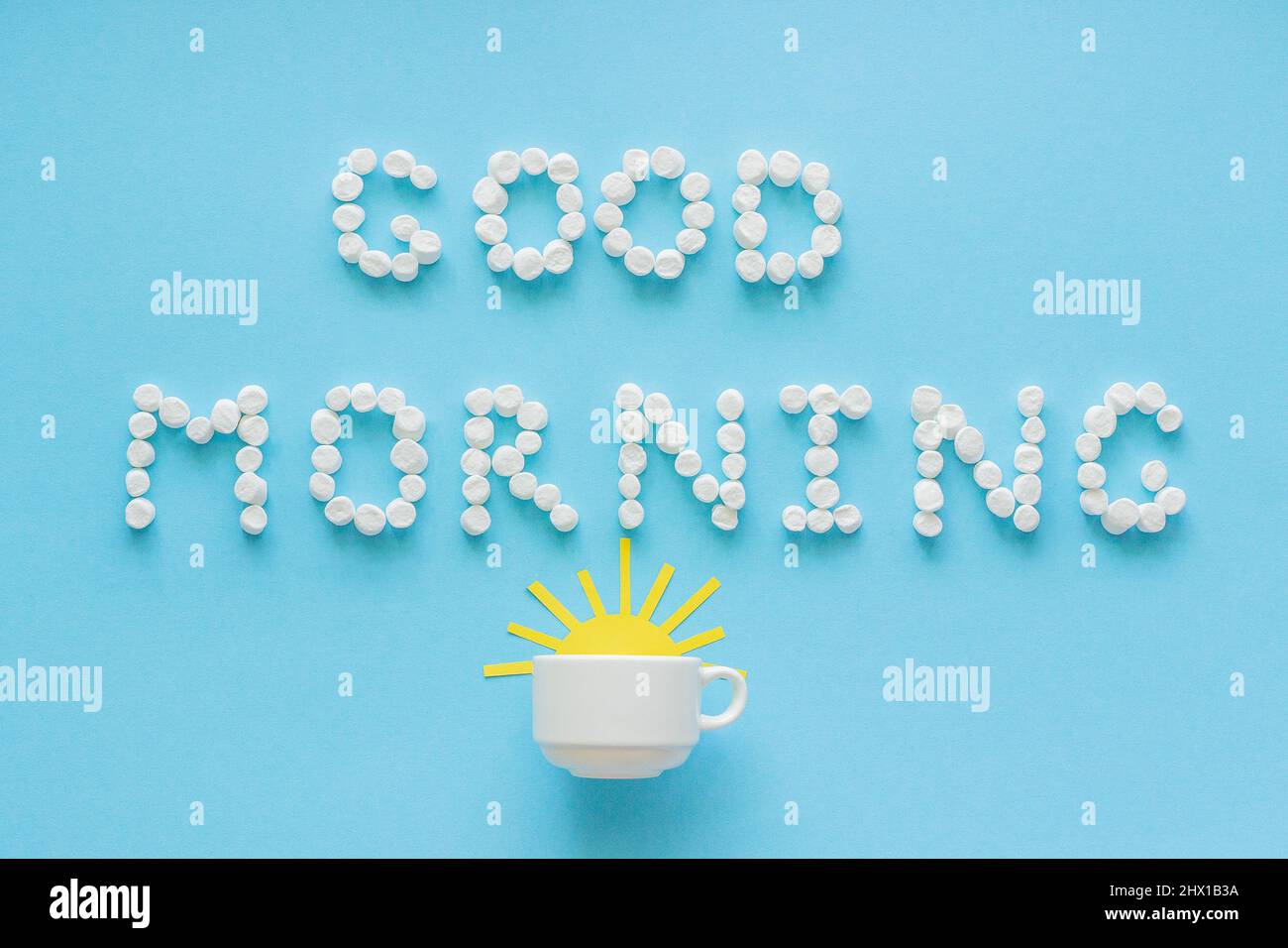 Testo Buongiorno da marshmallow e tazza di caffè con giallo sorgere sole su sfondo blu. Biglietto d'auguri Concept Top View Foto Stock