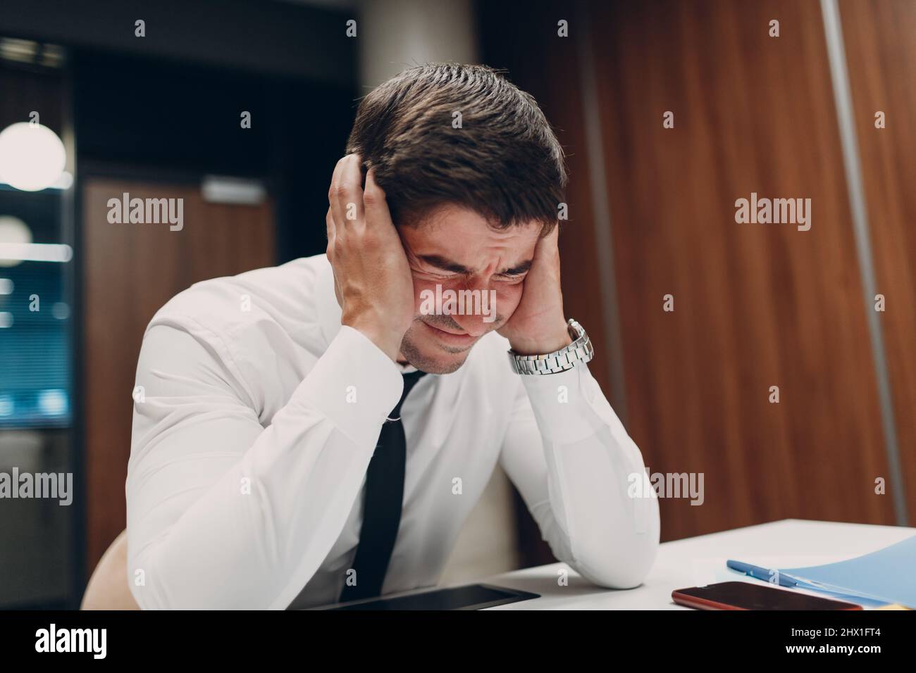 Triste giovane uomo d'affari siediti e tieni la testa con le mani al tavolo d'ufficio. Concetto di sanzioni economiche, crisi finanziaria e fallimento Foto Stock