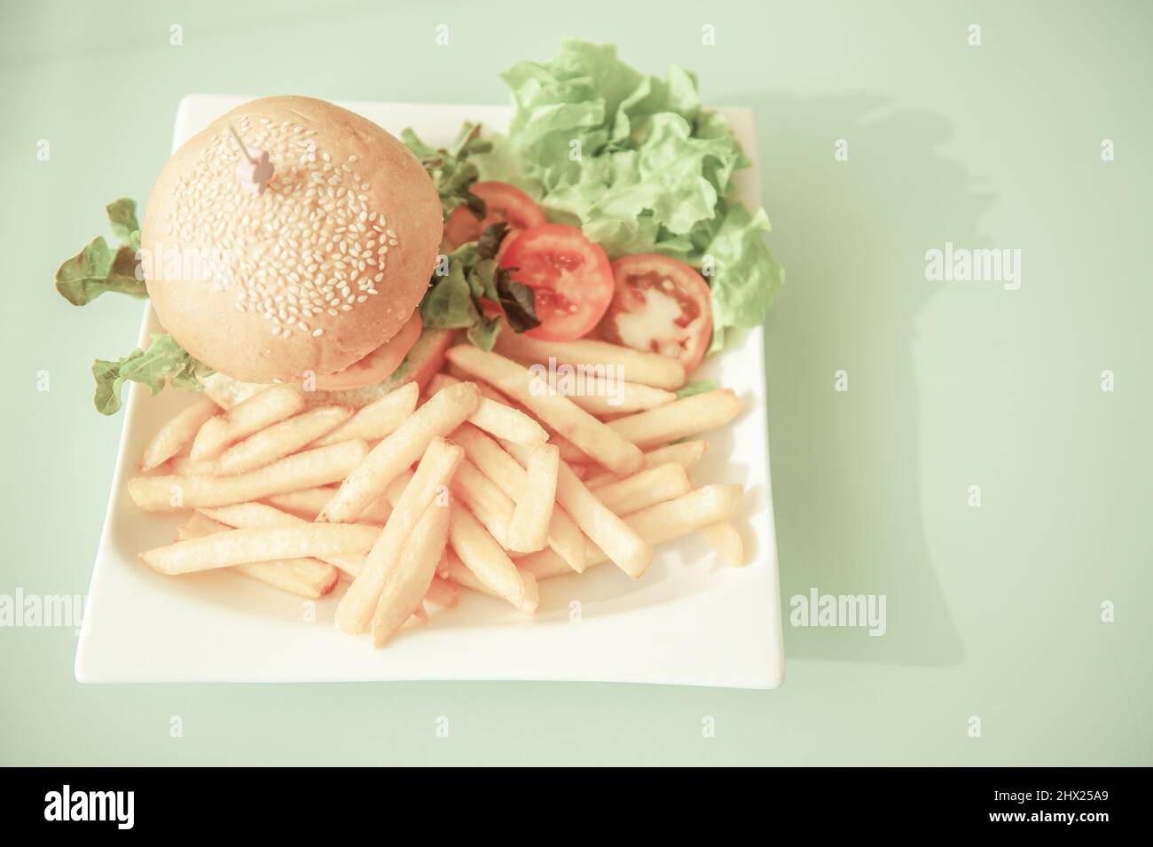 Hamburger con sesamo e fritte francesi con verdure servite su un piatto bianco su un tavolo verde chiaro sullo sfondo. Vista dall'alto. Disposizione piatta. Primo piano. Foto Stock