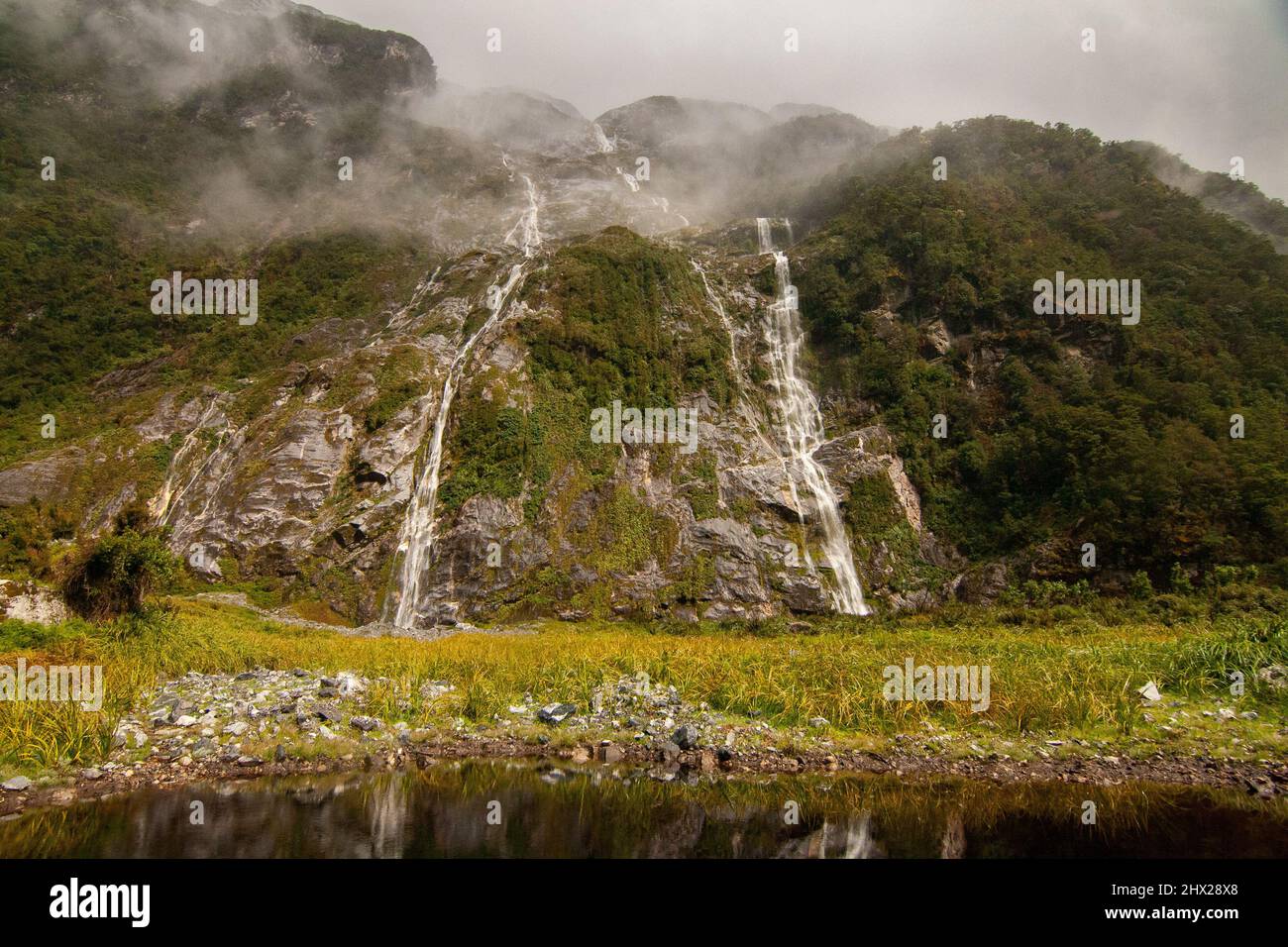 Specchio cascata del lago. Le montagne di Fiordland scure e foggiose cadono in acqua e il lago nero, Milford Track Nuova Zelanda Foto Stock