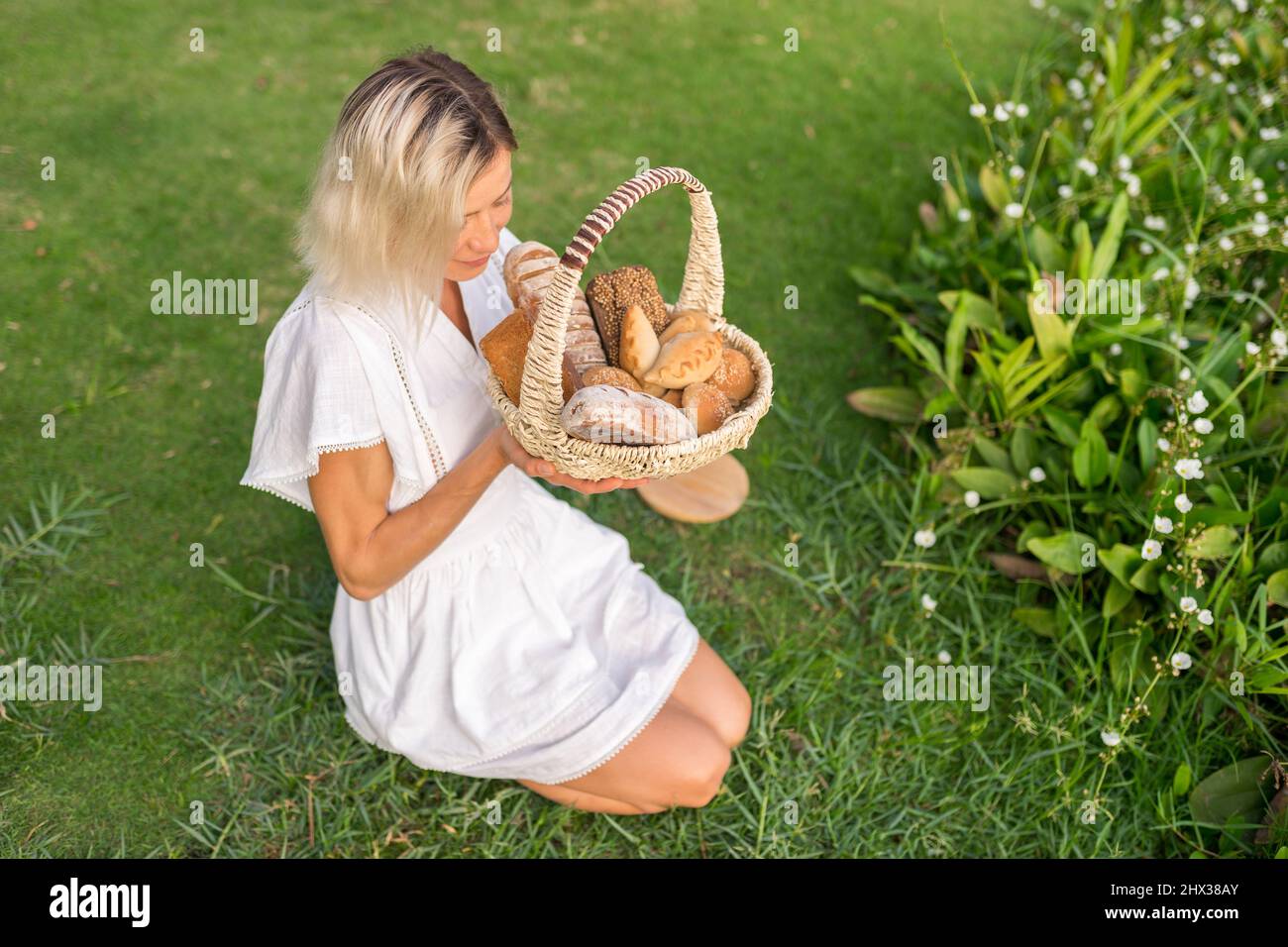 Giovane donna in vestito bianco seduta sull'erba con cestino con assorti di pane casereccio marrone e bianco. Foto di alta qualità Foto Stock