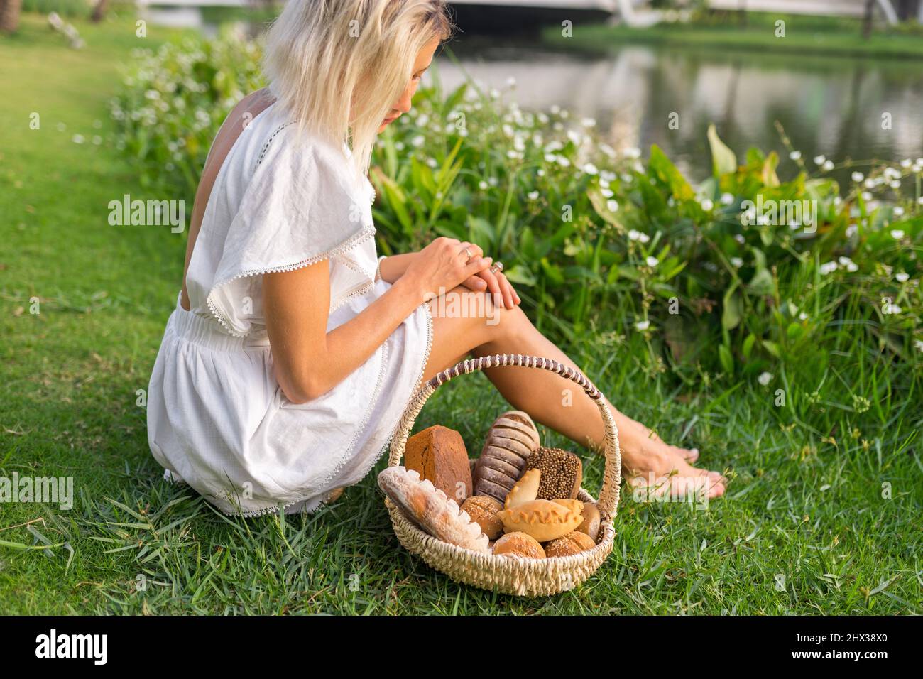Giovane donna in vestito bianco seduta sull'erba con cestino con assorti di pane casereccio marrone e bianco. Foto di alta qualità Foto Stock