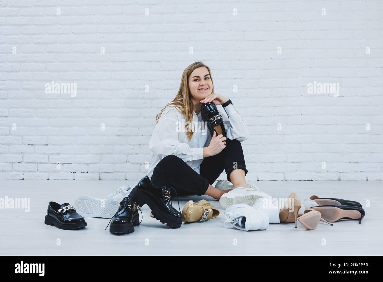 Una giovane donna si siede sul pavimento tra le scarpe e sceglie nuove  scarpe per se stessa. Donna in una camicia bianca e pantaloni neri. Donna  in scarpe bianche Foto stock -