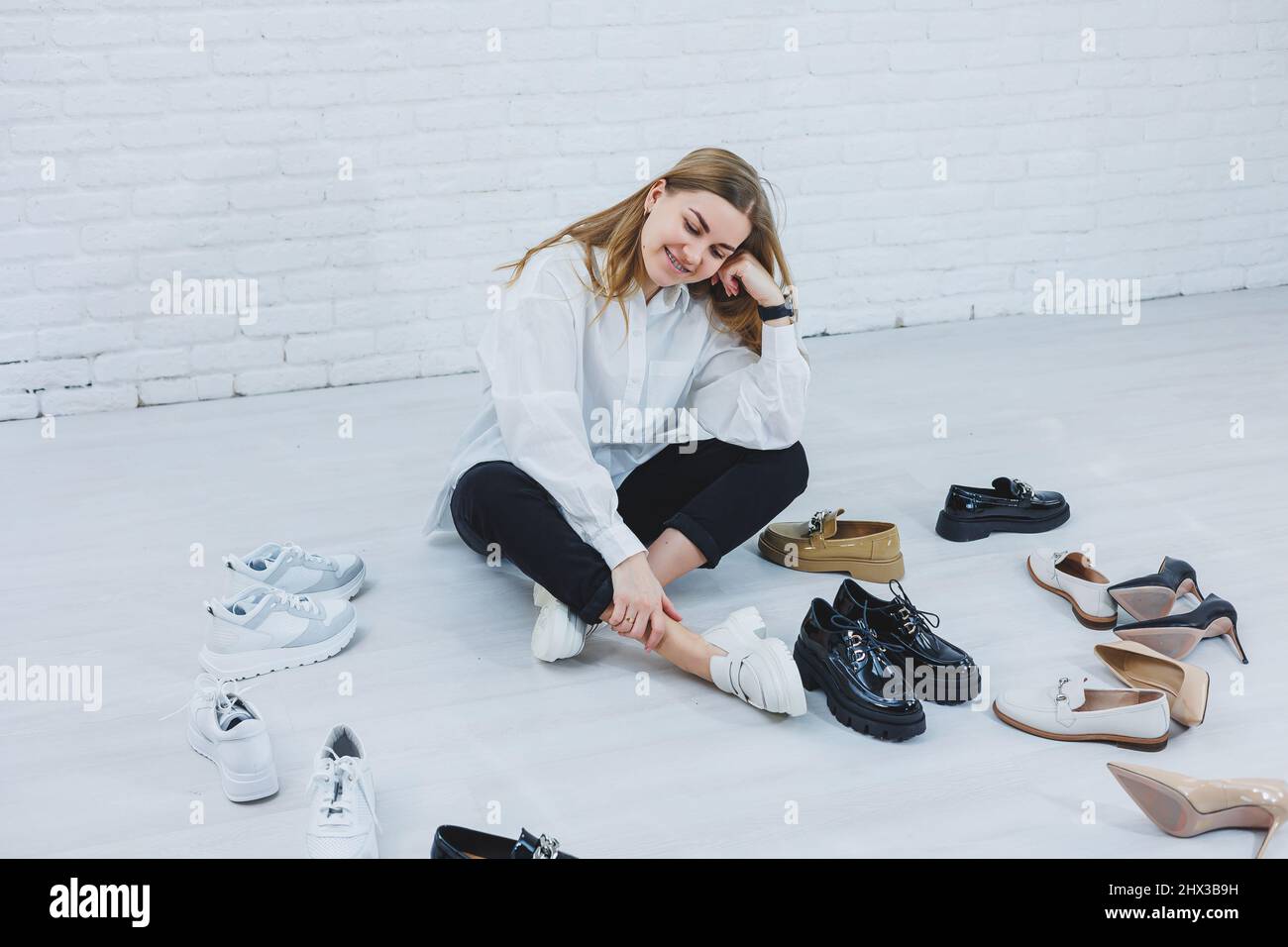 Una giovane donna si siede sul pavimento tra le scarpe e sceglie nuove  scarpe per se stessa. Donna in una camicia bianca e pantaloni neri. Donna  in scarpe bianche Foto stock -