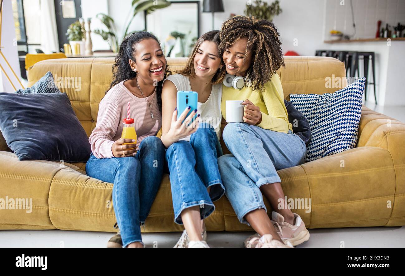 Tre giovani donne multirazziali che prendono selfie mentre si rilassano insieme a casa Foto Stock