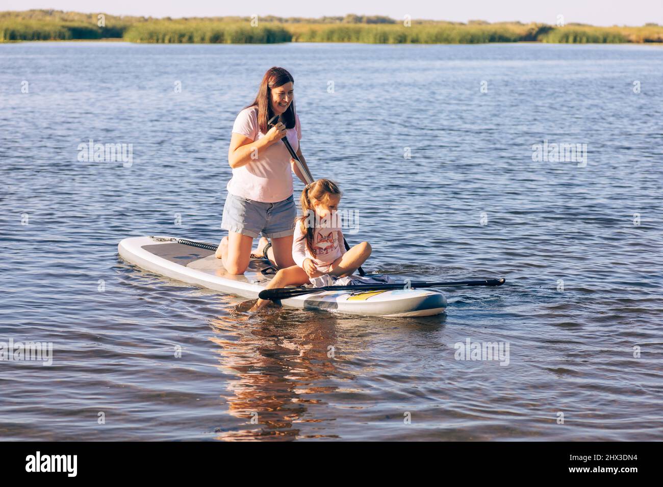 Madre caucasica di mezza età con la bambina sup a bordo sia sorridente che guardando giù l'acqua. Stile di vita attivo e tempo di famiglia insieme Foto Stock