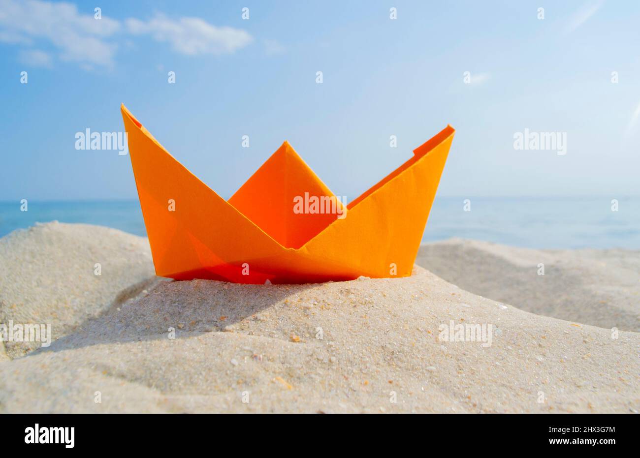 Barca di carta arancione brillante in sabbia gialla sullo sfondo delle onde del mare e del cielo blu Foto Stock