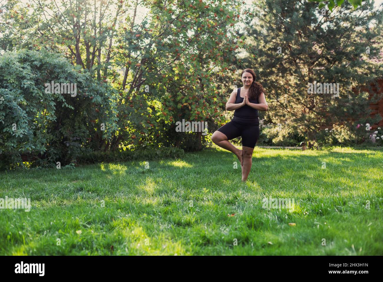 Femmina con peso extra in piedi su un piede in posizione yoga godendo il momento su erba verde sul cortile di cottage con casa di legno e alberi Foto Stock