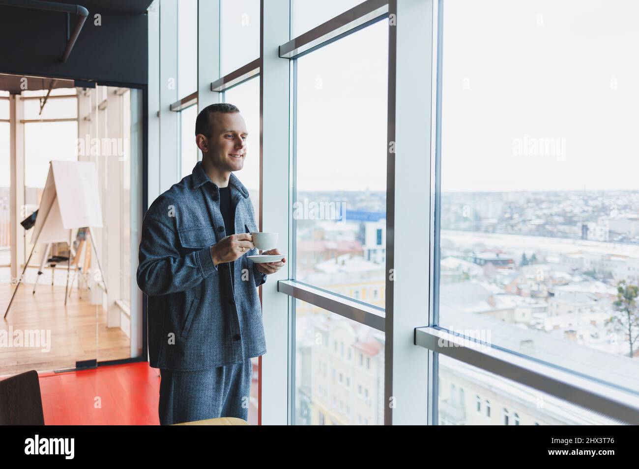 Un giovane di aspetto europeo in un abito informale beve caffè e guarda fuori dalla finestra Foto Stock