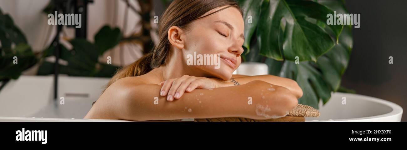 Giovane donna sognante appoggiata sul lato della vasca da bagno e riposata nel bagno decorato con piante tropicali Foto Stock