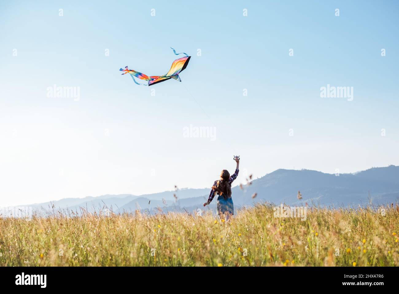 Ragazza dai capelli lunghi con volare un aquilone colorato sul prato d'erba alta nei campi di montagna. Momenti felici dell'infanzia o concetto di tempo libero all'aperto Foto Stock