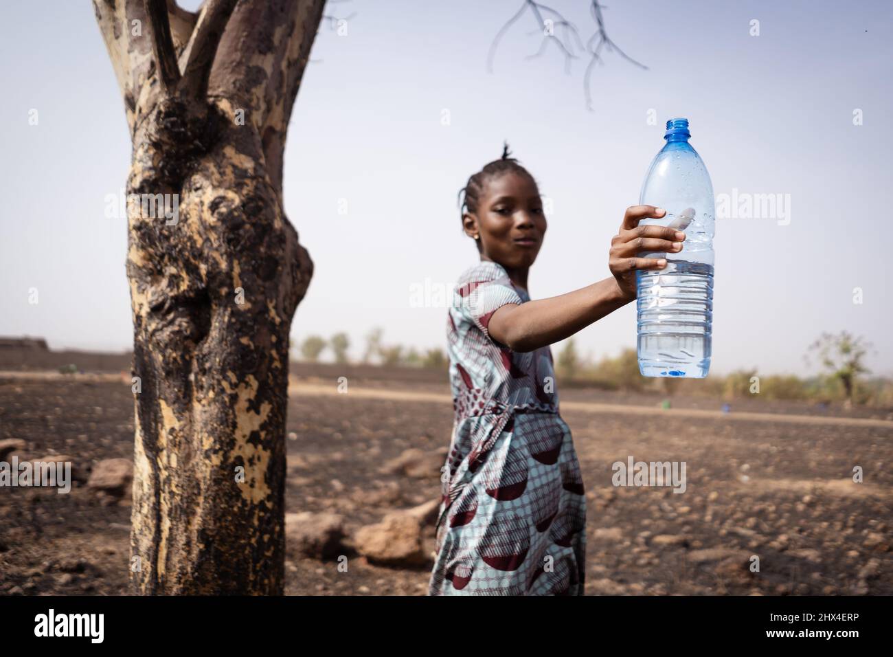 Carina ragazza di villaggio africano con un boccale grande di acqua nella sua bocca e tenendo aloft una bottiglia di plastica, raccomandando l'uso di drinki pulito e fresco Foto Stock