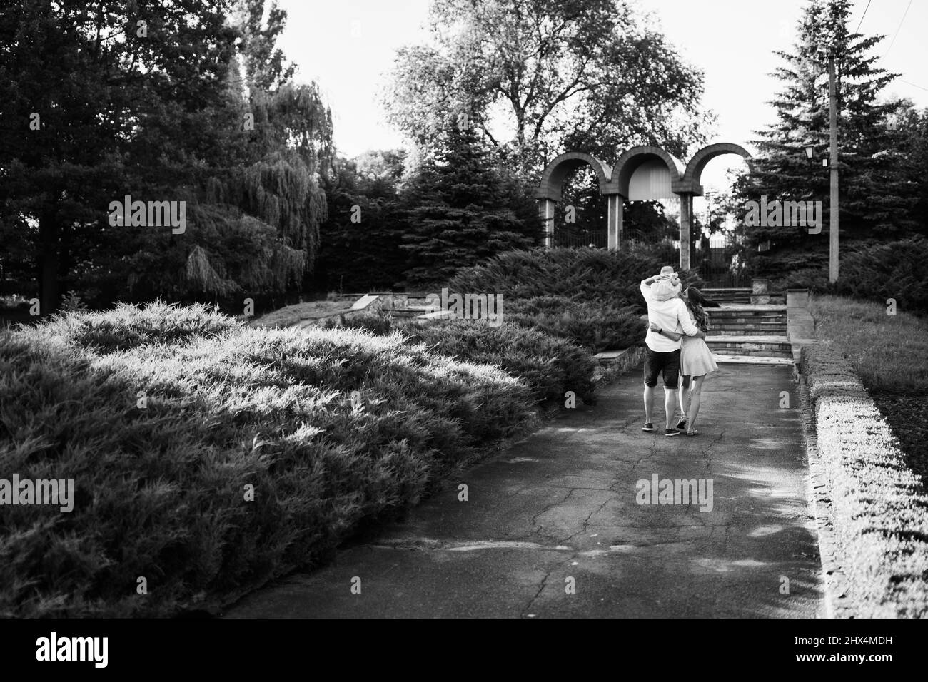 Moglie amorevole e marito premuroso che abbraccia il parco, giovani genitori con bambina a piedi, sorridente, simpatico bambino seduto sulla spalla dei padri, cal Foto Stock