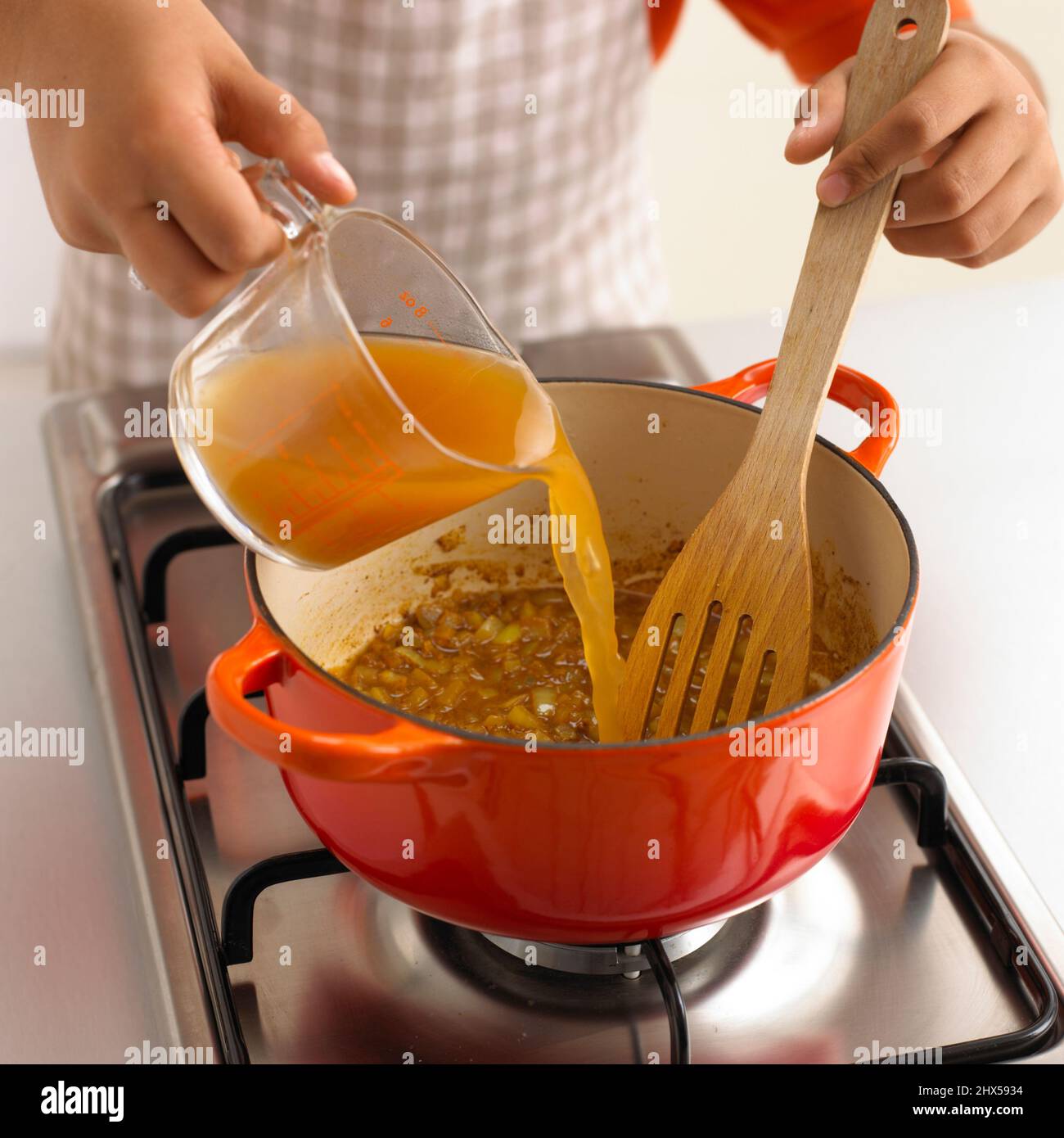 Ragazzo versare il brodo dalla caraffa di vetro agli ingredienti in una casseruola sul piano cottura a gas, primo piano Foto Stock