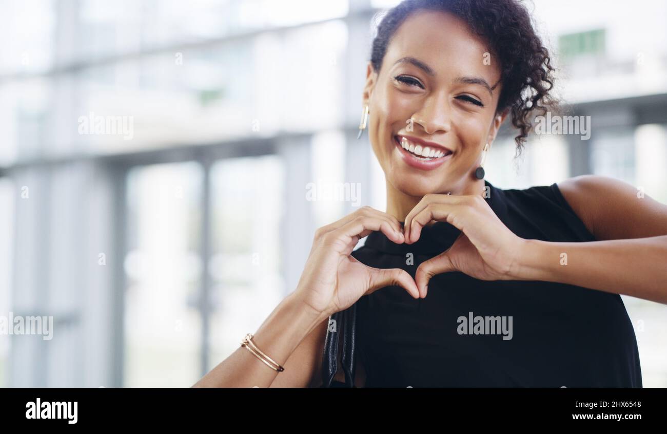 Fare ciò che amo e amare ciò che faccio. Scatto corto di una giovane donna d'affari che mostra un segno del cuore mentre cammina attraverso un ufficio moderno. Foto Stock