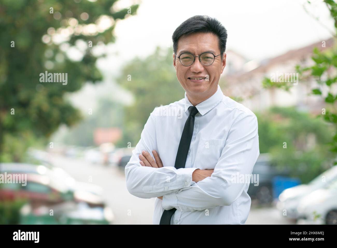 Ben vestito uomo d'affari asiatico maturo con le braccia incrociate, sorridendo con fiducia. Proprietà residenziale urbana sullo sfondo. Foto Stock