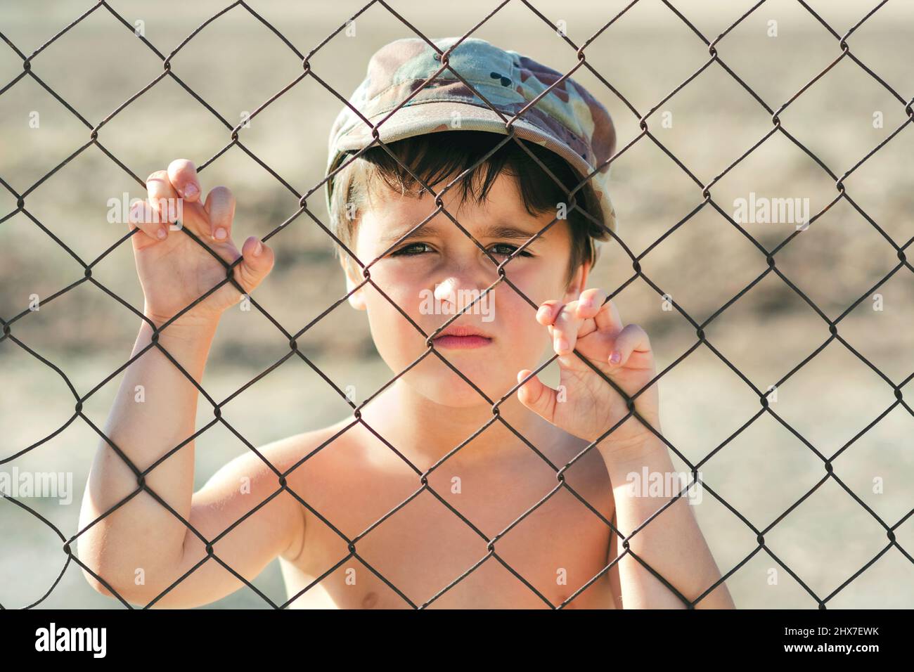 Bambino triste in un tetto militare dietro una recinzione. Arrestare il concetto di guerra Foto Stock