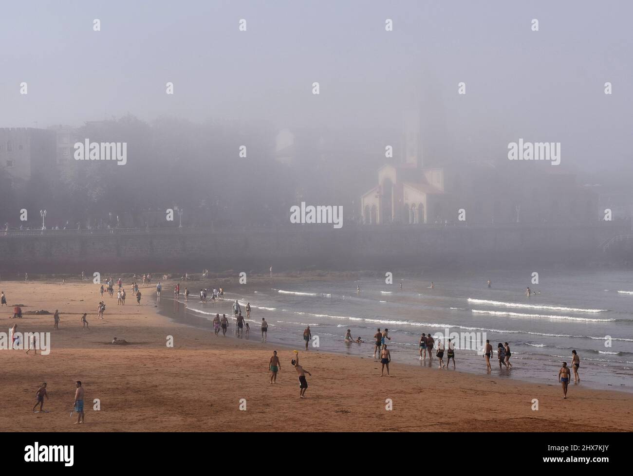 Gente vacanzieri sulla spiaggia di Playa de San Lorenzo con la chiesa di San Pedro in background. Pomeriggio estivo di nebbia a Gijon Asturias Spagna. Foto Stock