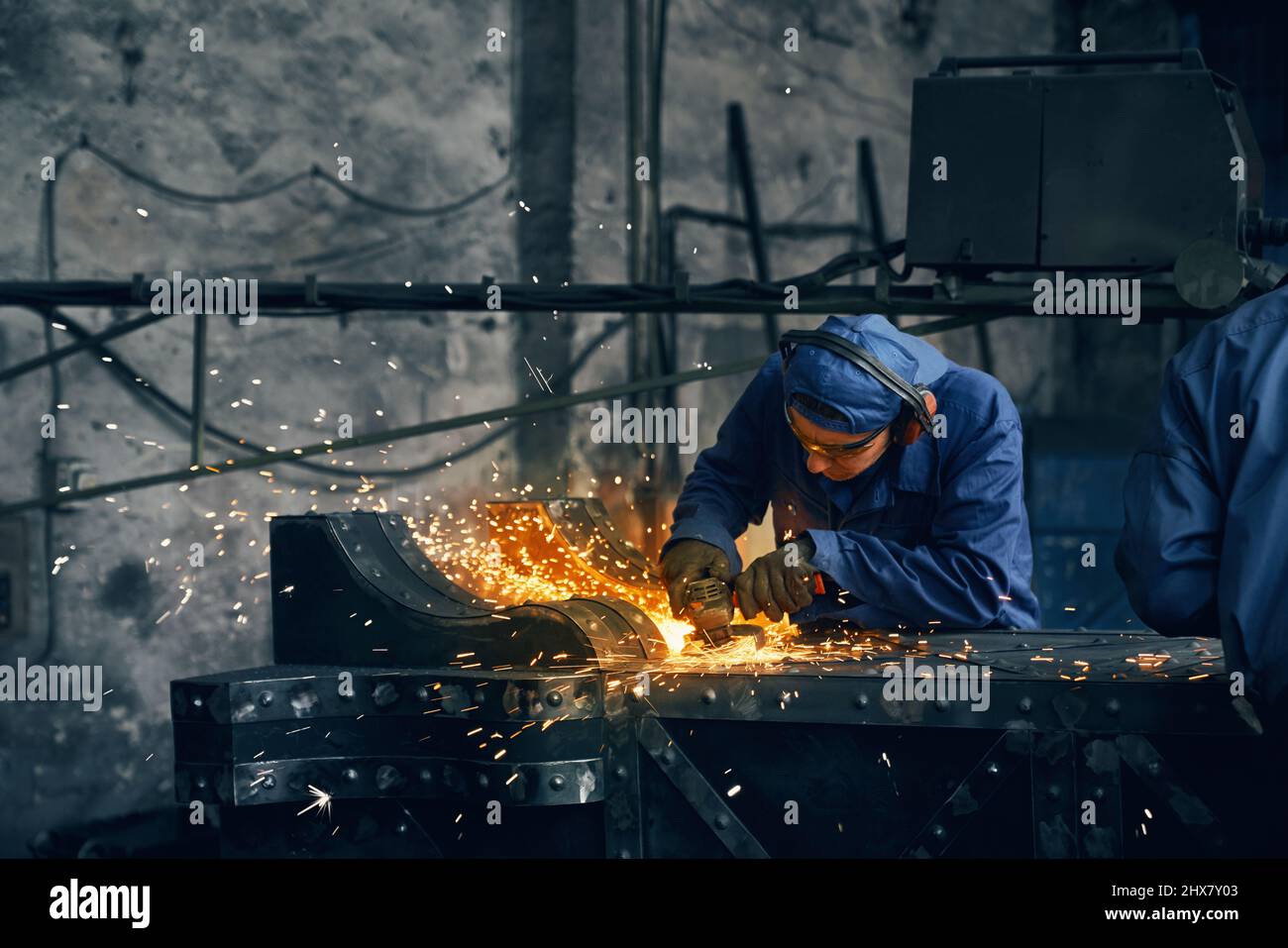 Primo piano dell'uomo in speciale uniforme blu scuro che fa cancelli da ferro in garage o officina. Concetto di lavorazione con rettificatrice in garage. Foto Stock