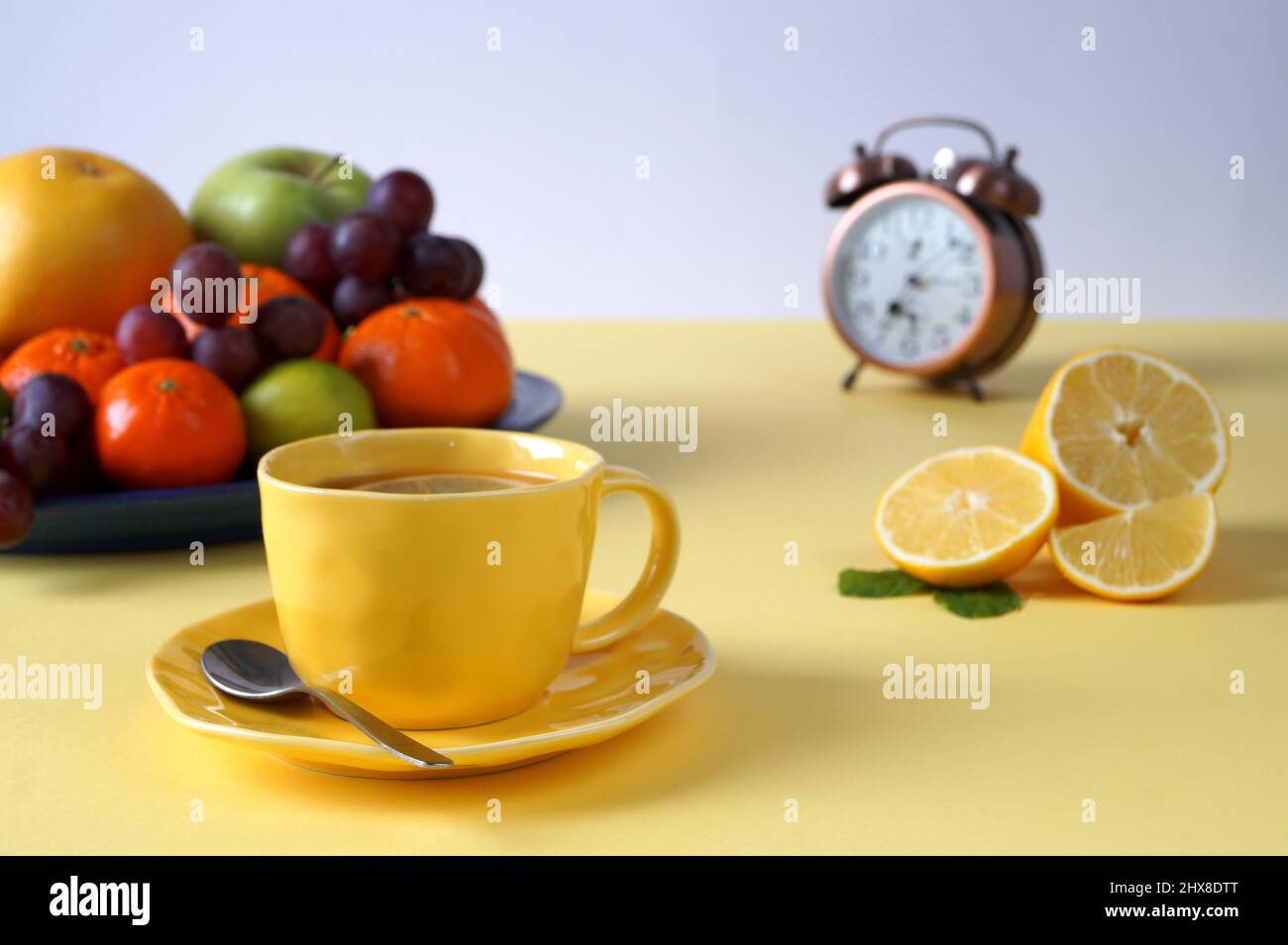 Sana colazione di frutta e tè con limone. C'è una tovaglia gialla sul tavolo, una tazza di tè è gialla. È presente una sveglia sfocata Foto Stock