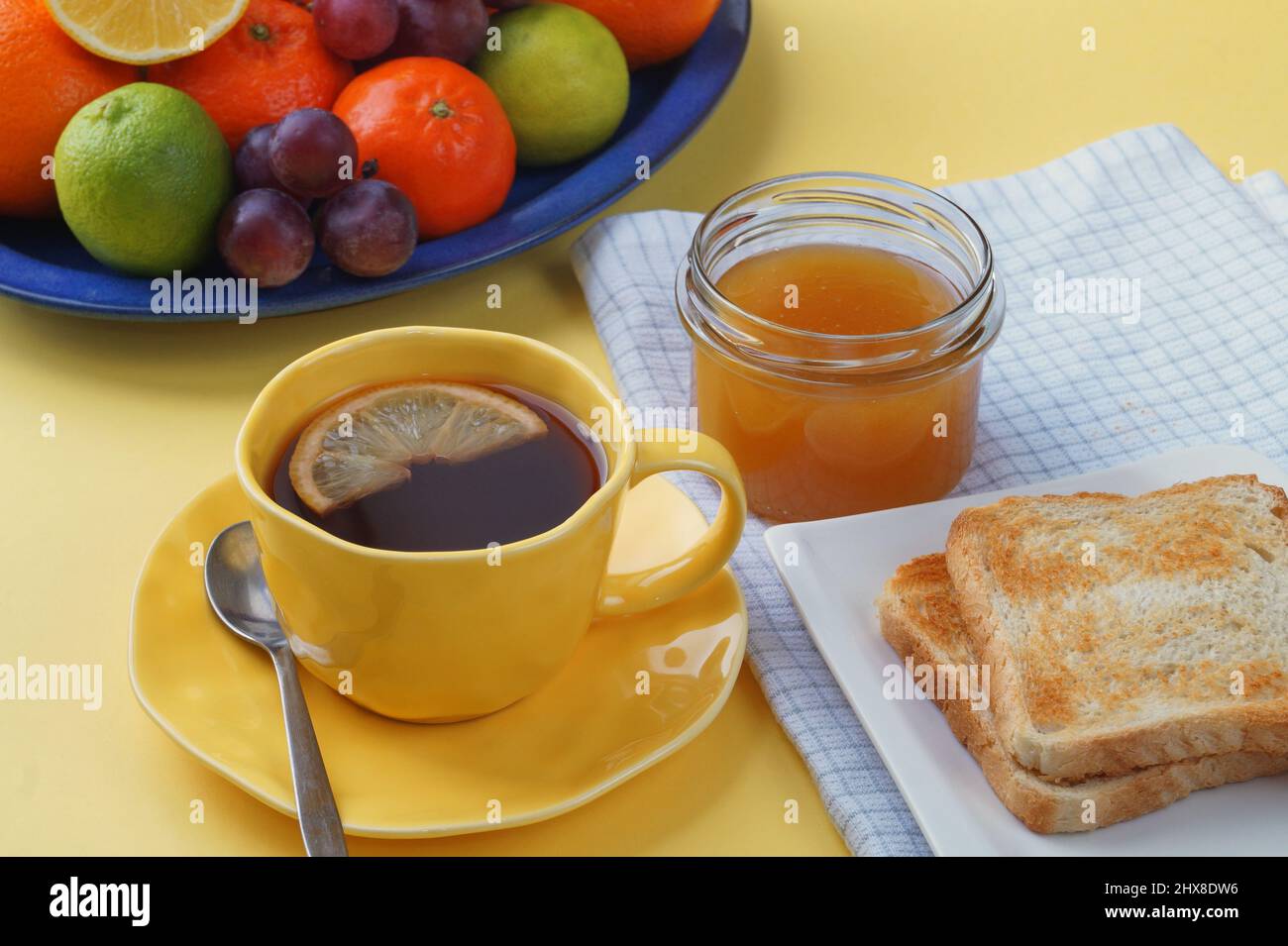 Colazione - pane tostato, tè con limone, miele in un vaso, frutta su un piatto. E una bella tovaglia gialla. Foto Stock
