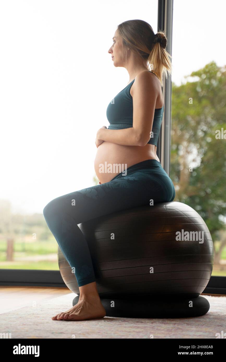 Donna incinta bionda che pratica yoga con una grande palla a casa Foto Stock