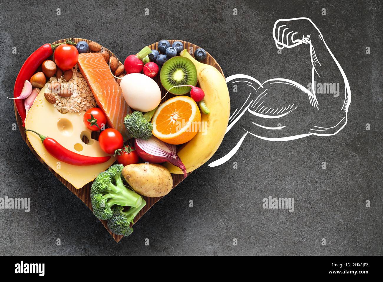 Una dieta sana ed equilibrata. Cibo sano su un tagliere di legno a forma di cuore e un braccio muscolare come sinonimo di forza e salute. Foto Stock