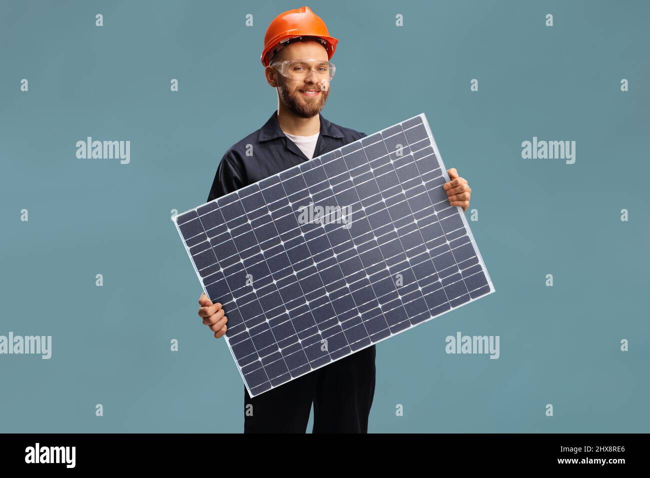 Giovane lavoratore maschile in un unibody che tiene un modulo fotovoltaico a celle solari isolato su sfondo blu Foto Stock