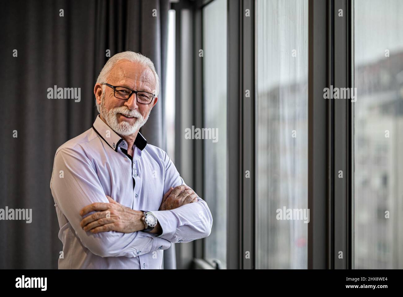 Ritratto di sorridente allegro soddisfatto maturo bianco barbuto uomo con occhiali in piedi accanto alla grande finestra nel suo ufficio braccia incrociate guardando ca Foto Stock