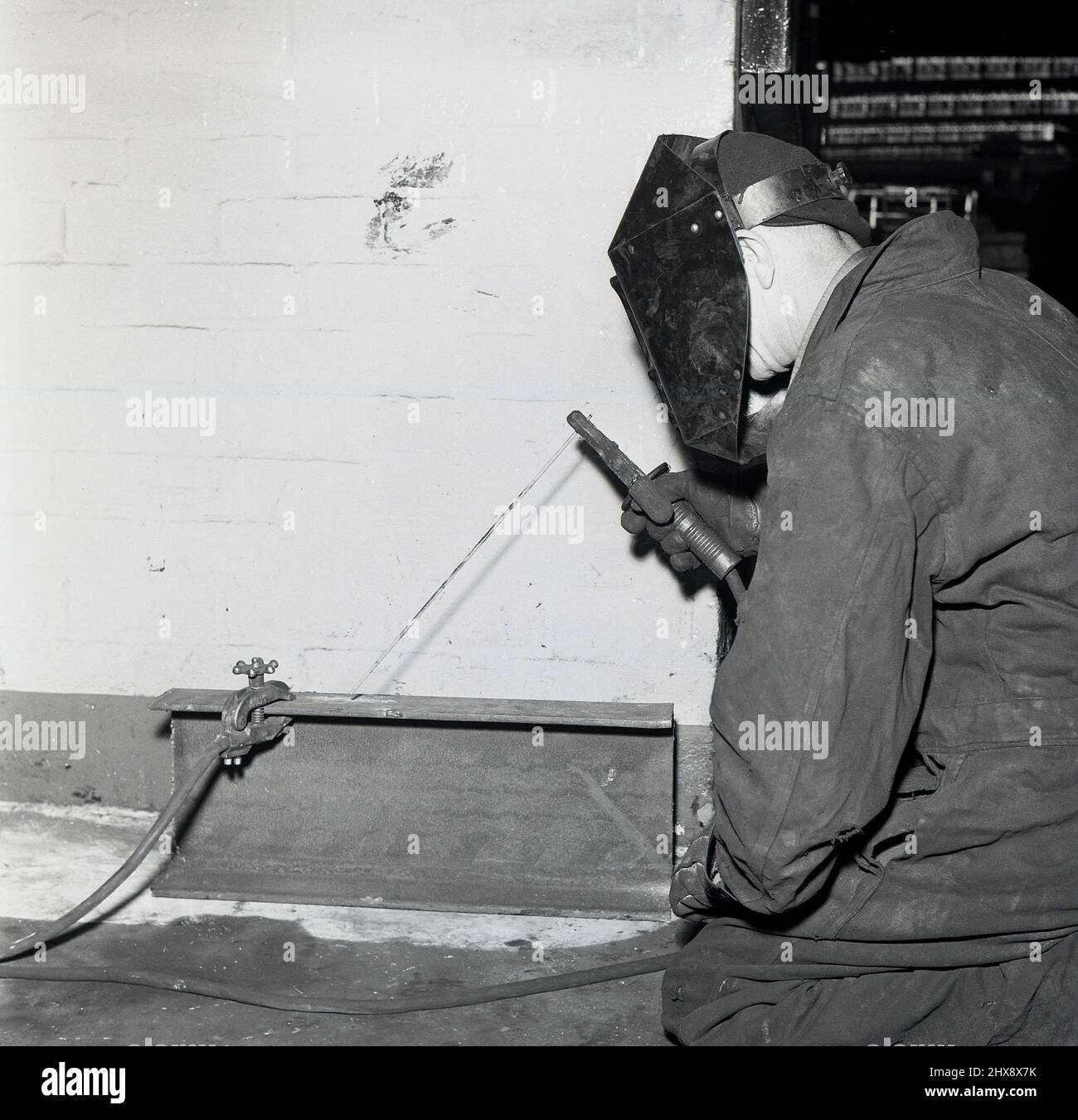 1950s, storico, un lavoratore che indossa una maschera o uno schermo protettivo per impieghi pesanti e guanti che utilizza una saldatrice ad arco elettrica su una piastra metallica su una piccola trave in acciaio, Abbey Works, Port Talbot, Neath, Galles, REGNO UNITO. Foto Stock