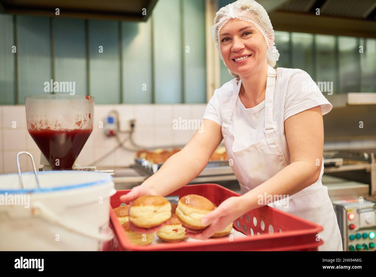 Giovane donna come panettiere che riempie le frittelle berlinesi con marmellata nel panificio Foto Stock