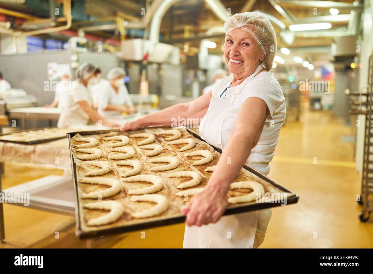 La panetteria femminile anziana porta una teglia da forno con molti croissant per la cottura nella panetteria di grandi dimensioni Foto Stock