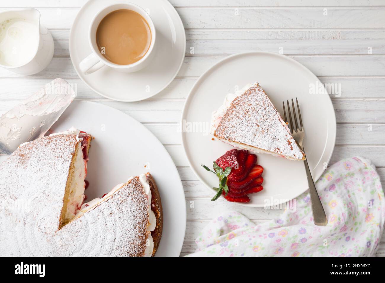 Vista dall'alto di un pan di Spagna Vittoria fatto in casa riempito con fragole, marmellata e panna montata su un tavolo di legno bianco e una tazza di caffè o tè Foto Stock