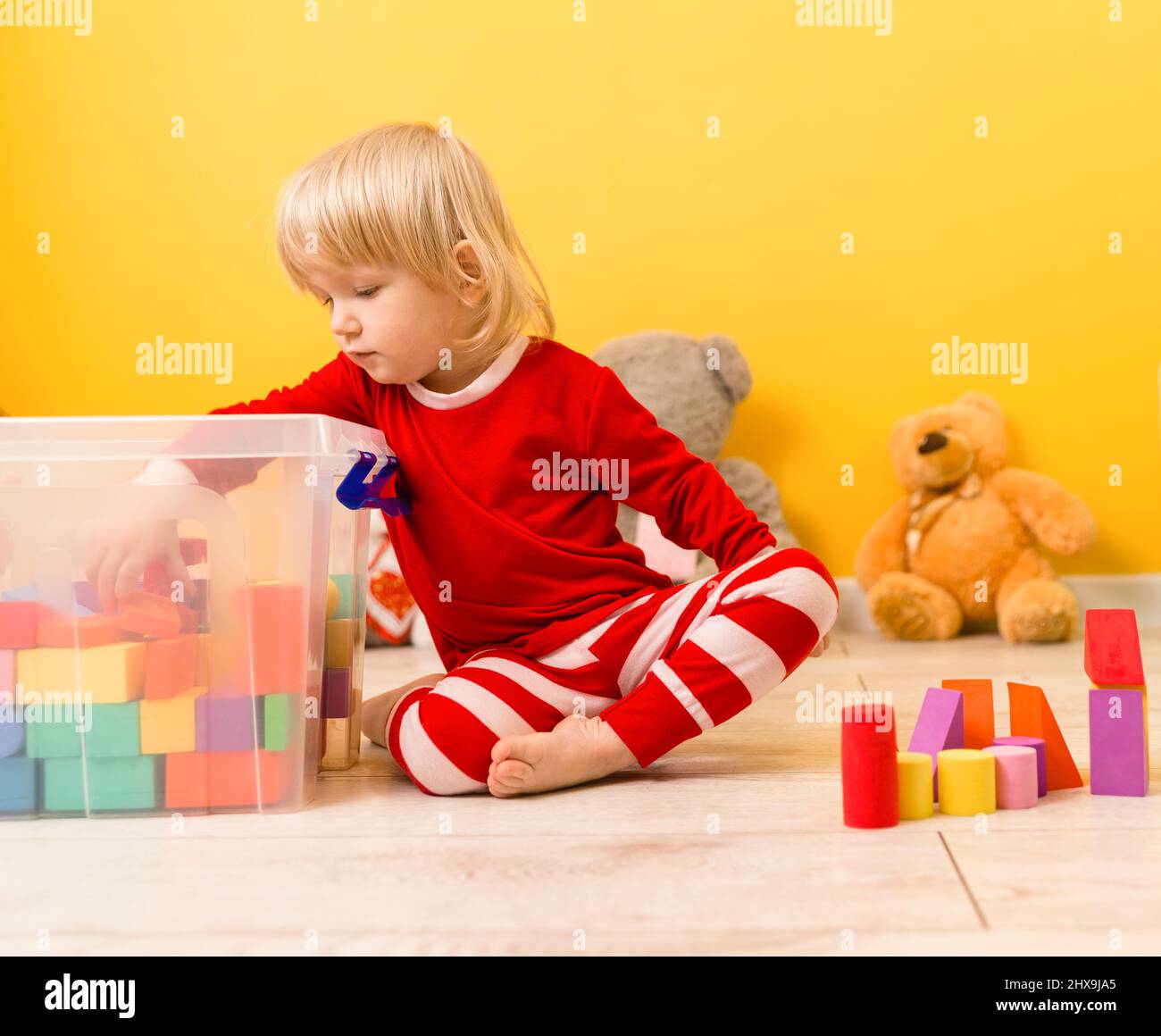 Un bambino bello in pigiama rossa si siede sul pavimento nella sala giochi ed elimina i cubi colorati da una scatola trasparente. Messa a fuoco selettiva Foto Stock