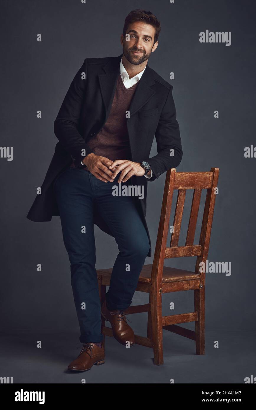 Fai il tuo segno con stile. Ritratto di studio di un giovane uomo vestito con eleganza in piedi accanto ad una sedia su sfondo grigio. Foto Stock