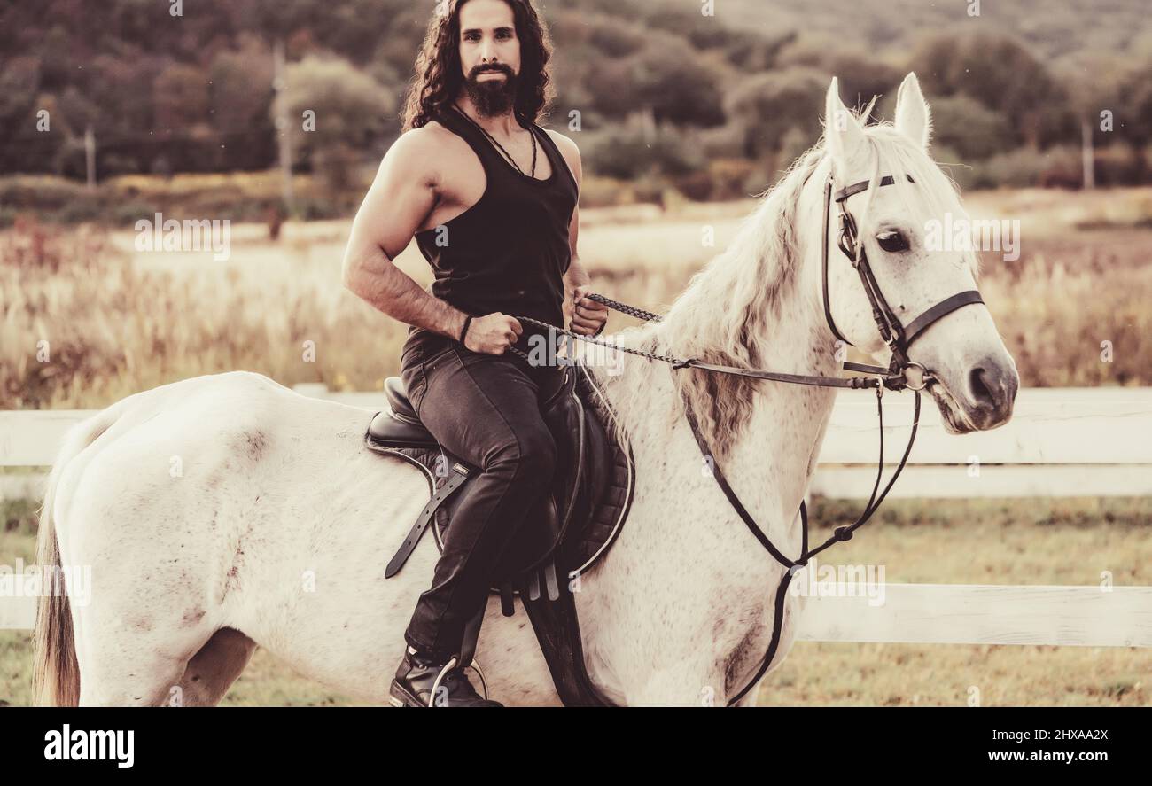 Giovane jockey allenando il suo cavallo per un giro. Bella vista, hunky cowboy cavalca a cavallo in bella natura lato campagna. L'equestre cavalca il suo pozzo bianco Foto Stock