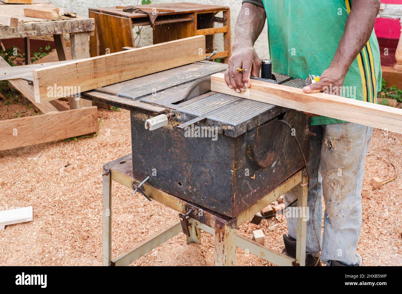 Un costruttore di cabinate con un pennel tra le sue dita sostiene un pezzo di legno come lui usa una vecchia sega elettrica per tagliare la sua estremità Foto Stock