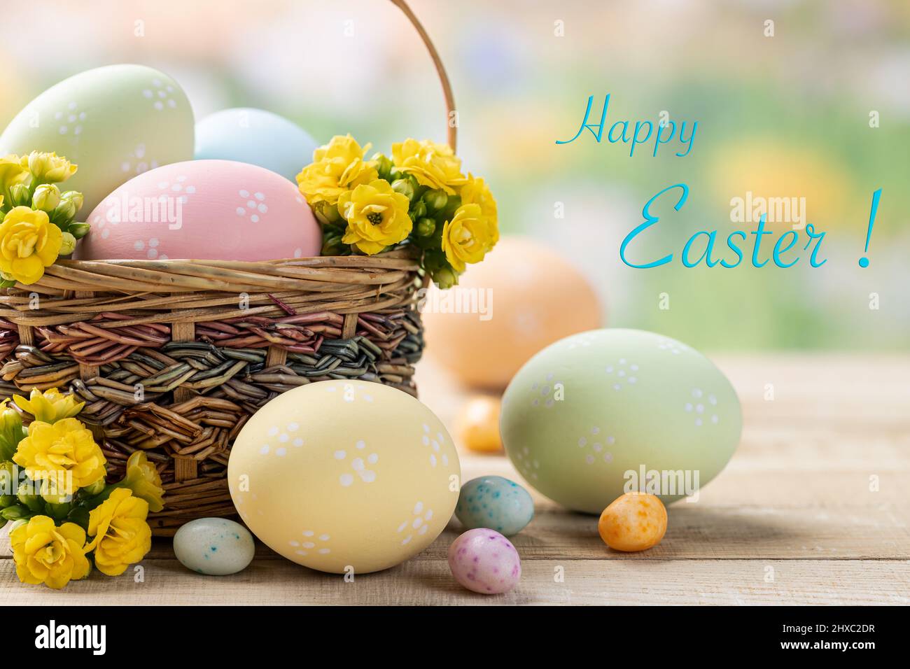 Uova e fiori di Pasqua in un cestino e su un tavolo di legno con sfondo colorato e felice Pasqua testo Foto Stock