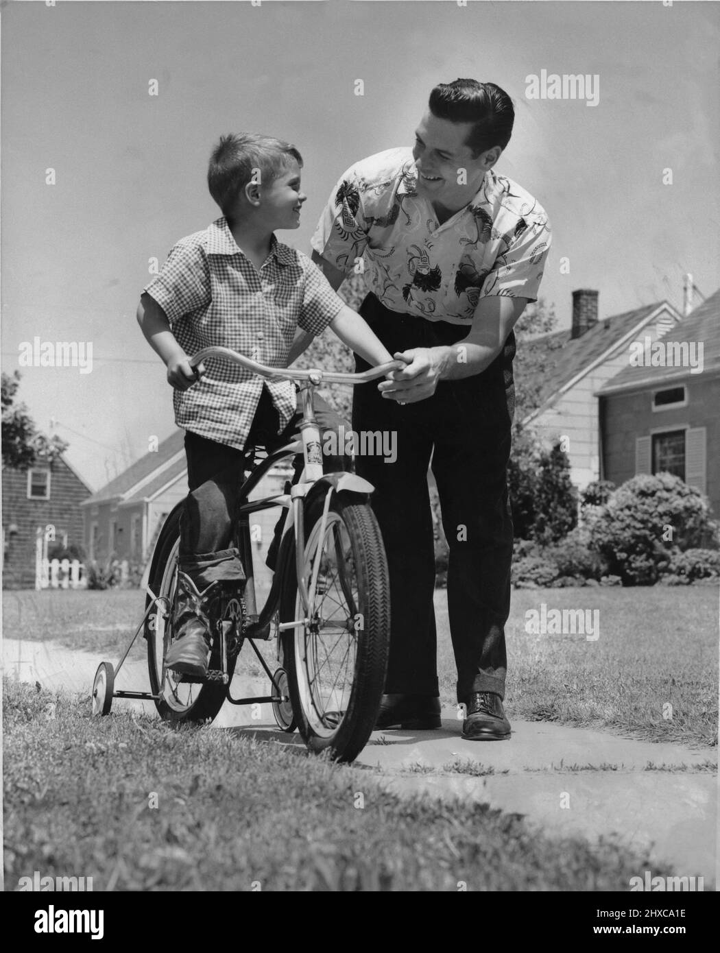 Papà e figlio si fermino per guardarci mentre imparano a fare un giro in bicicletta sul marciapiede in un quartiere residenziale Foto Stock