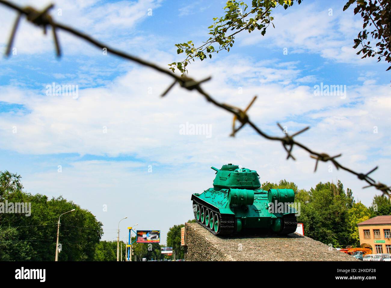 Monumento al vecchio serbatoio sovietico T-34. Un carro armato militare si erge su un piedistallo sullo sfondo di un cielo blu, filo spinato, alberi verdi, strada della città. Foto Stock