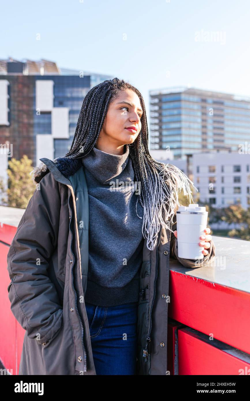 Sognante femmina ispanica con dreadlock in piedi vicino al confine con thermos bianco tazza di caffè caldo in città con edifici residenziali Foto Stock