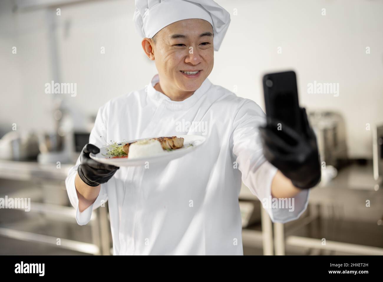 Chef asiatico che registra un vlog culinario al telefono, mostrando un piatto con pasto cotto in cucina. Concetto di video blogging e pasti di cottura per il ristorante Foto Stock