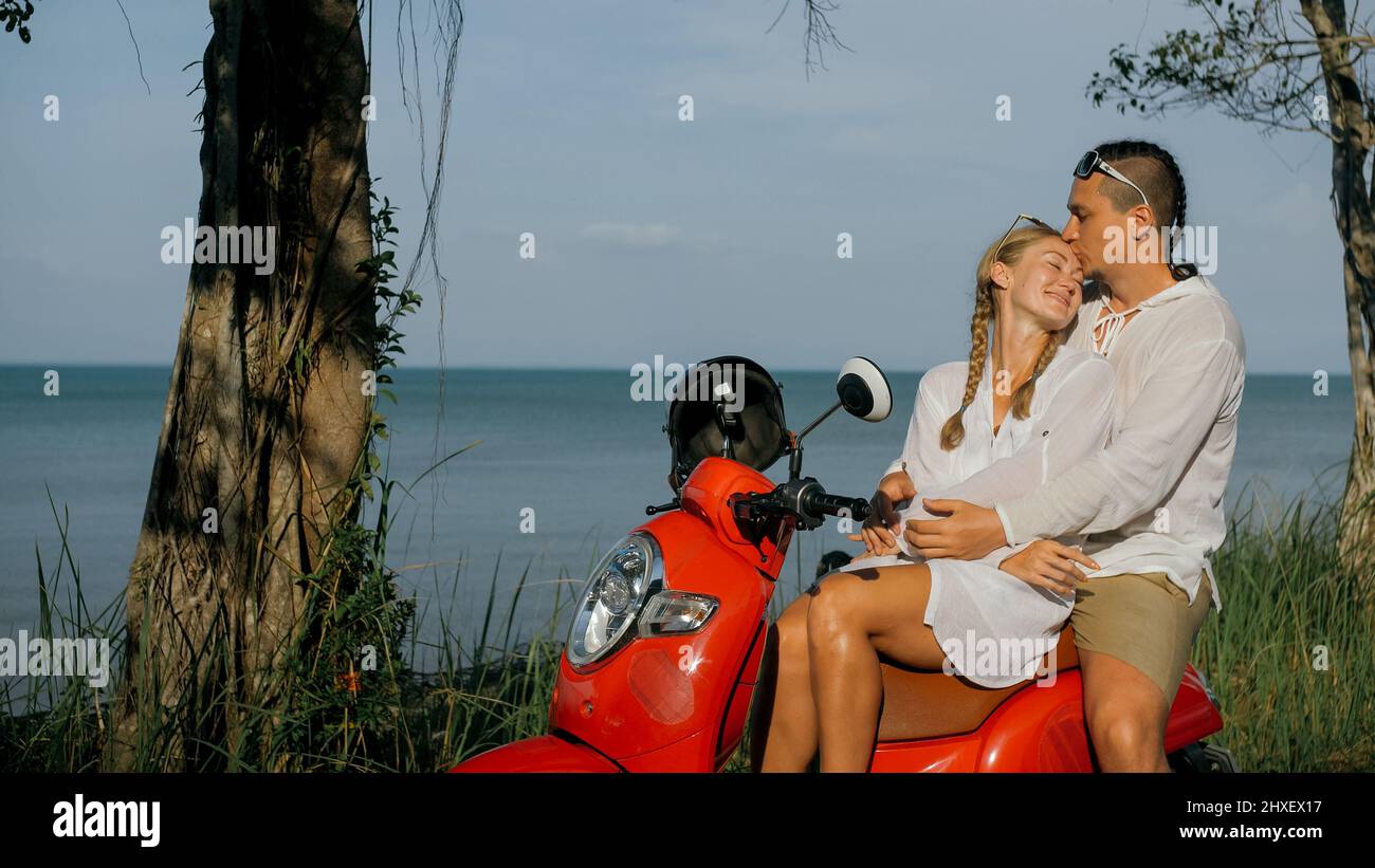 Viaggio su strada in scooter. Coppia d'amore su moto rossa in abiti bianchi sulla strada forestale. Appena sposato donna e uomo bacio, abbracci, sedersi in moto. Weddin Foto Stock