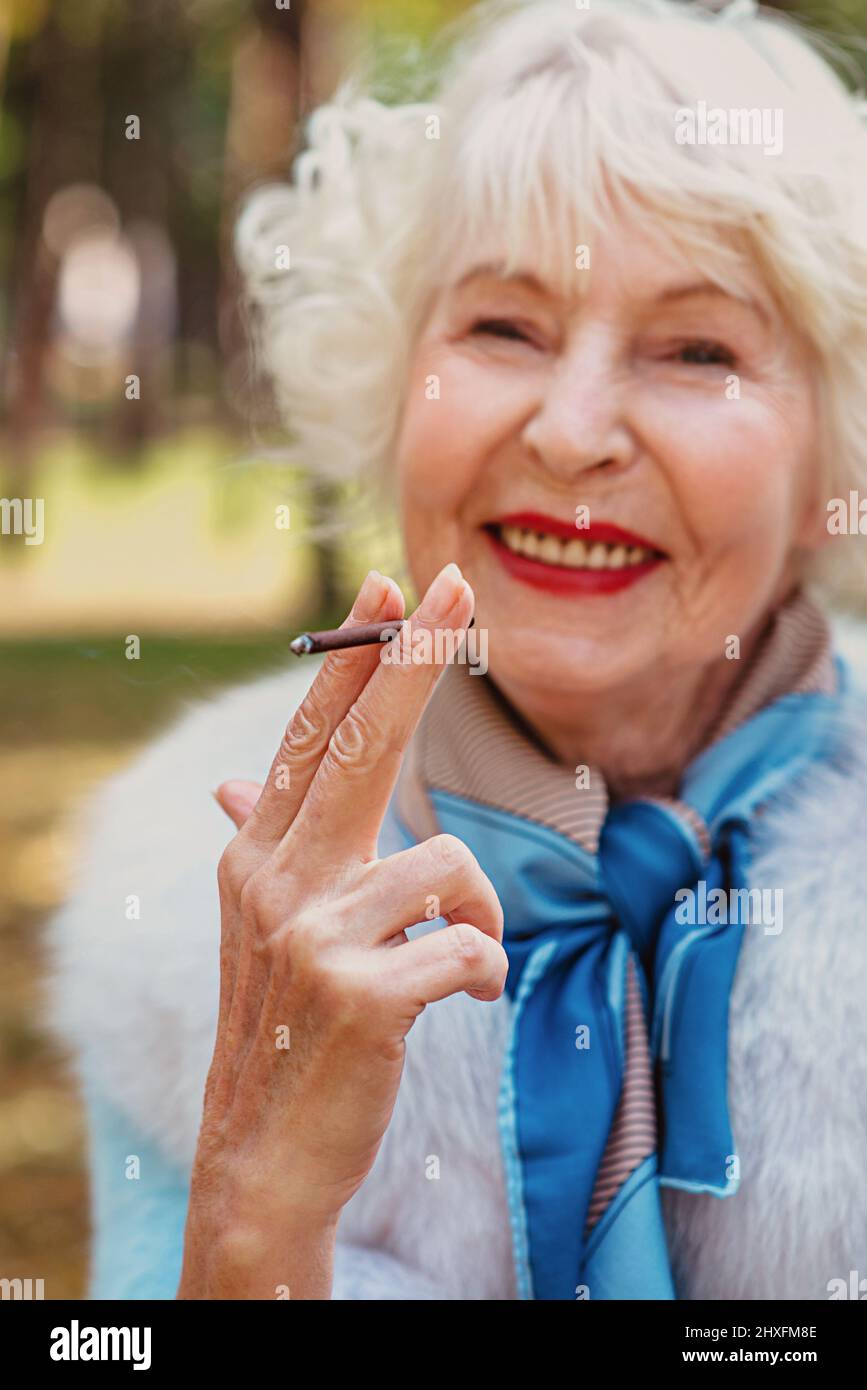 sorridente senior elegante donna alla moda con capelli grigi in pelliccia cappotto fumo sigaretta all'aperto. Stile di vita malsano, età, vibrazioni positive Foto Stock