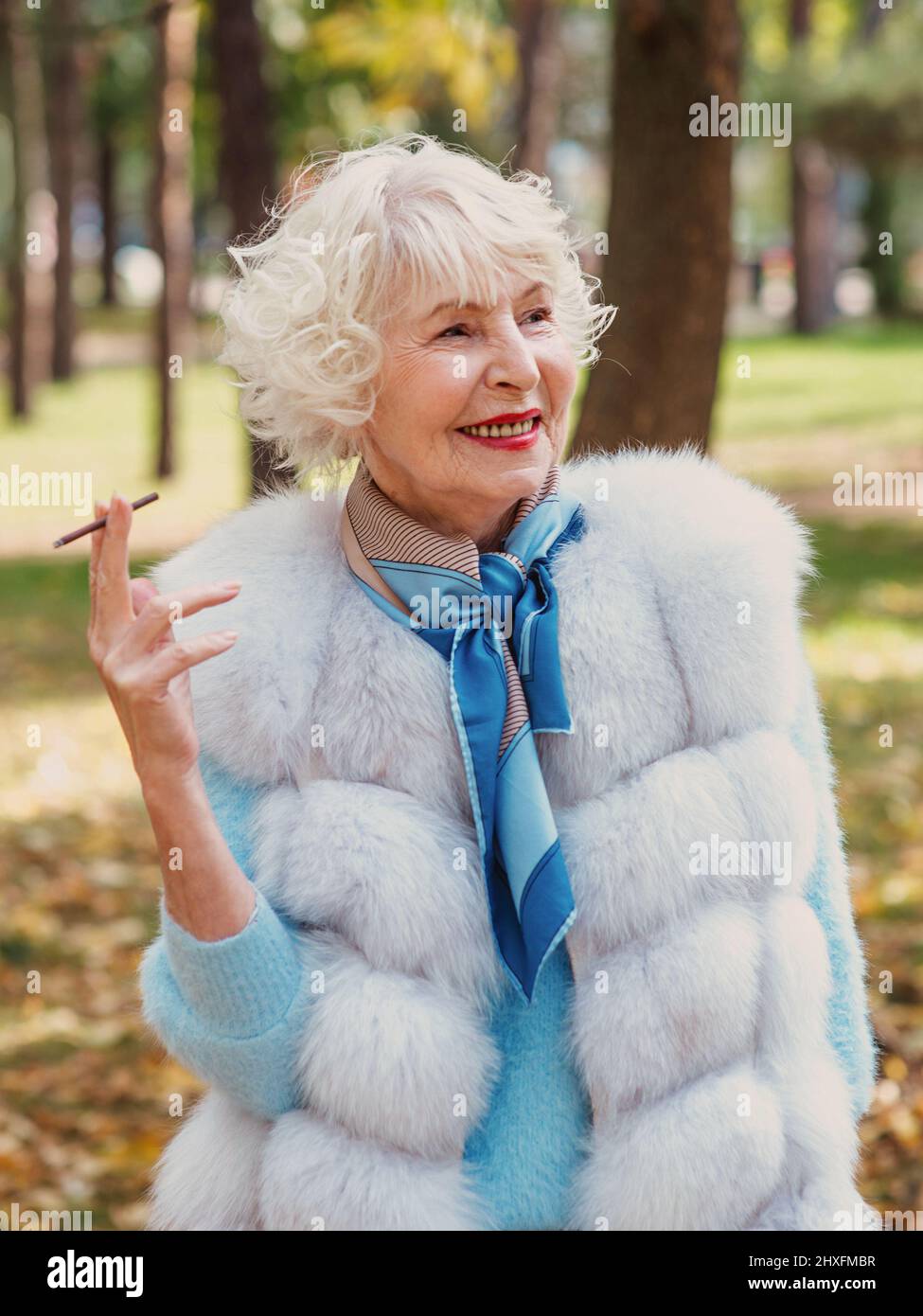 sorridente senior elegante donna alla moda con capelli grigi in pelliccia cappotto fumo sigaretta all'aperto. Stile di vita malsano, età, vibrazioni positive Foto Stock