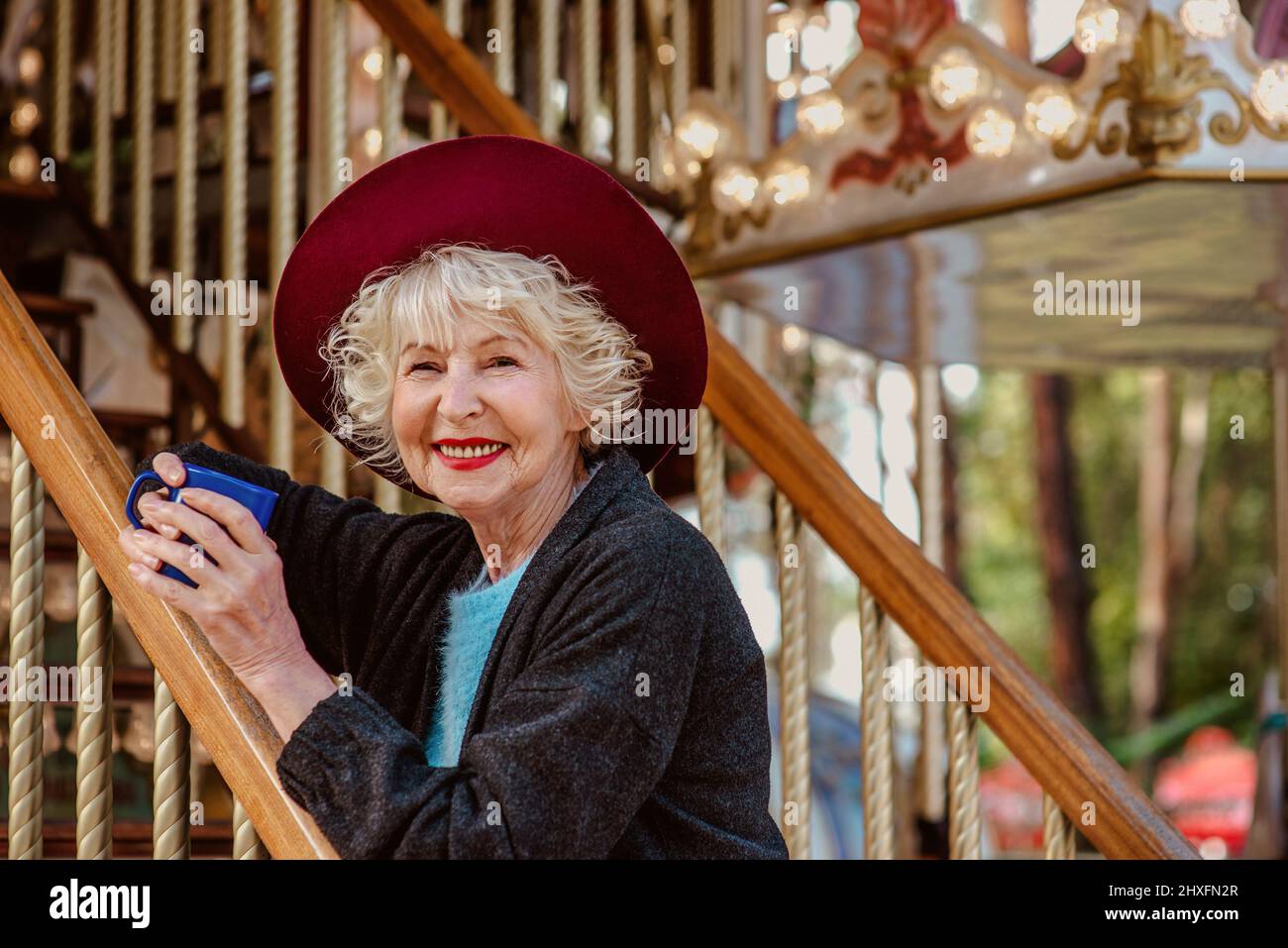 donna senior elegante in grigio scuro cappotto, cappello e con capelli grigi in piedi accanto alla giostra sorridente, bere caffè (tè) e godersi la vita. Foto Stock