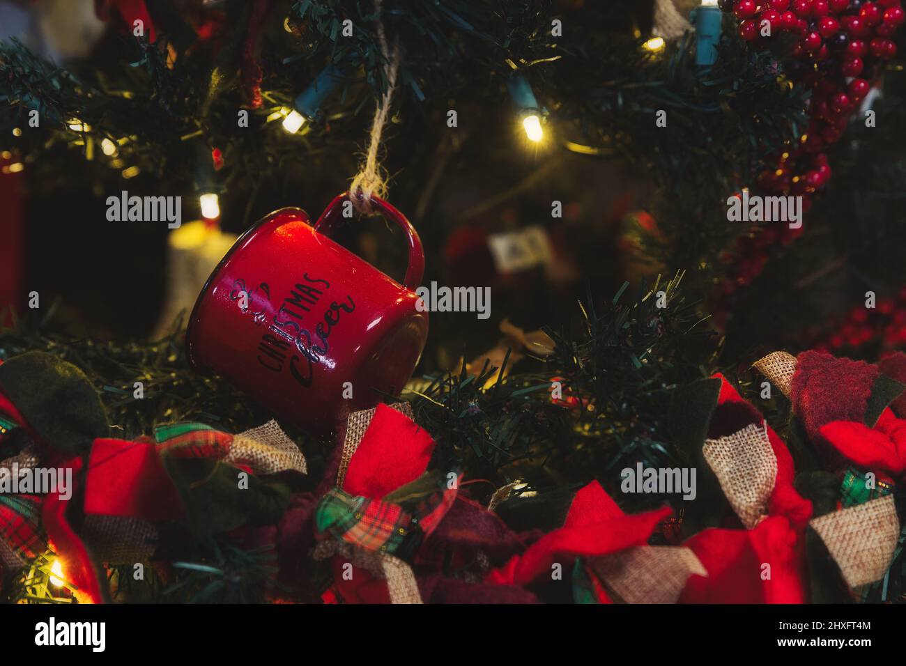 Un primo piano di una tazzina di lattina rossa appesa su un albero di Natale illuminato con ghirlanda di plaid Foto Stock