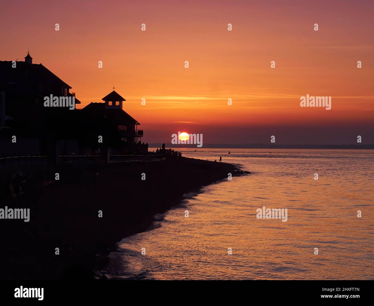 Un tramonto riccamente colorato visto da Cowes Beach, la luce del sole che passa attraverso le finestre di un edificio sul mare e le persone in silhouette sulla spiaggia. Foto Stock